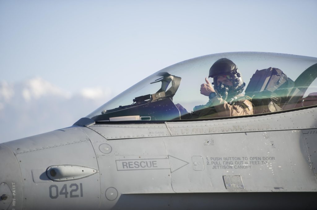 A view of a pilot in the cockpit of an American military jet. The pilot is giving the thumbs up sign with his left hand.